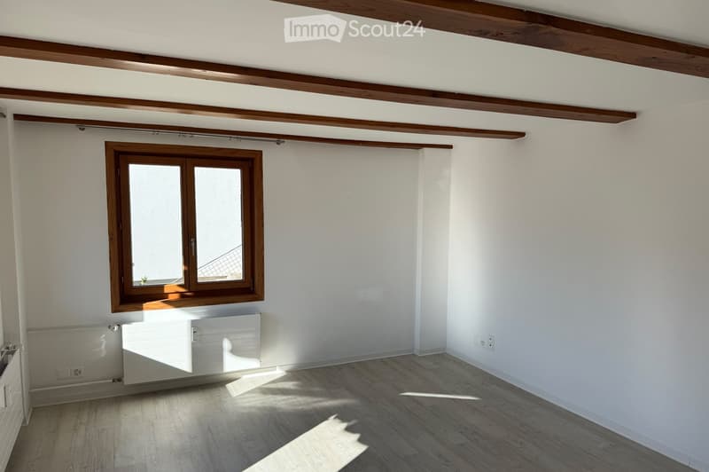 Empty room with wooden floor, white walls, exposed wooden beams on the ceiling, and a single window with wooden frame. No furniture or equipment present.