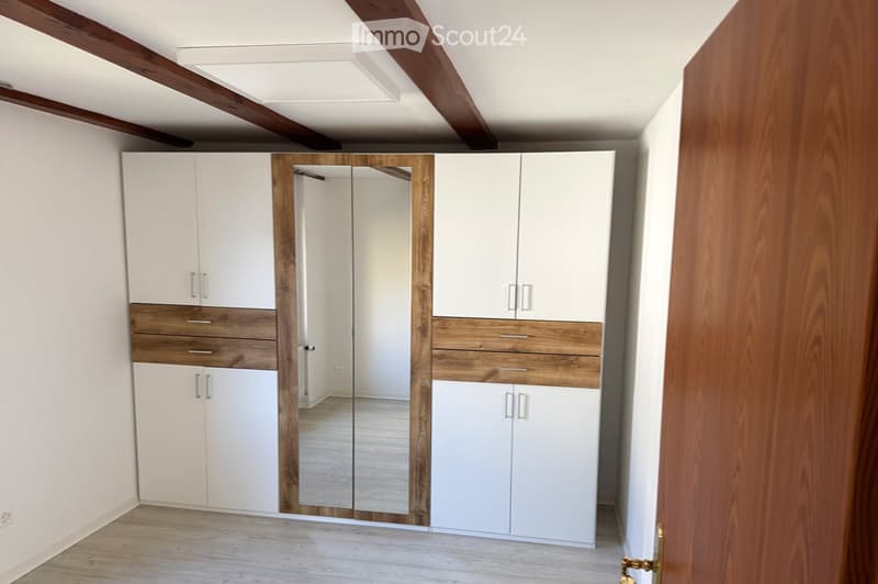 Empty room with white walls, wooden floor, and modern white wardrobes with wooden accents and a large mirror.