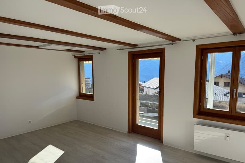Empty room with wooden flooring, white walls, exposed wooden beams on the ceiling, and large windows offering a scenic view of the mountains.