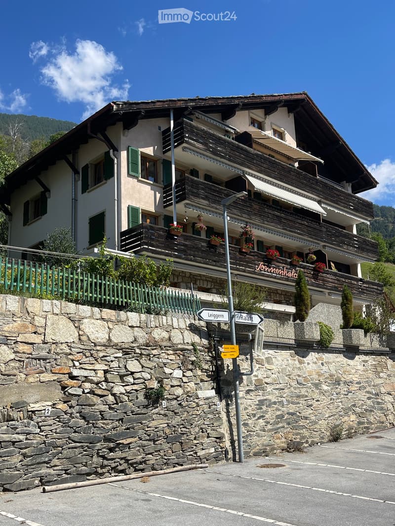 A multi-story building with a wooden balcony and green shutters, situated on a stone wall with street signs and a green fence.