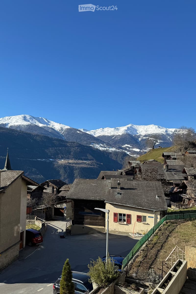 A picturesque village scene with traditional stone houses, red shutters, and a snow-capped mountain backdrop. The village features narrow streets, parked cars, and a small green fence along a hillside.