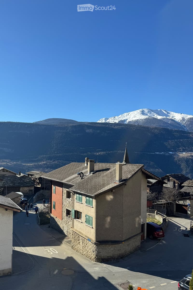 A multi-story building with a stone facade and green shutters, situated in a village with snow-capped mountains in the background.