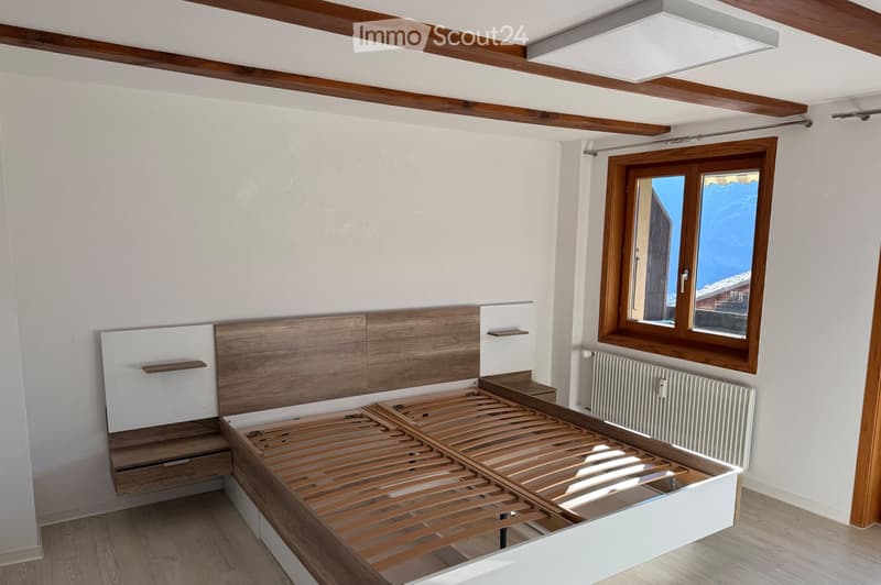Modern bedroom with a wooden bed frame, built-in storage, large window with wooden frame, radiator, and exposed wooden ceiling beams.