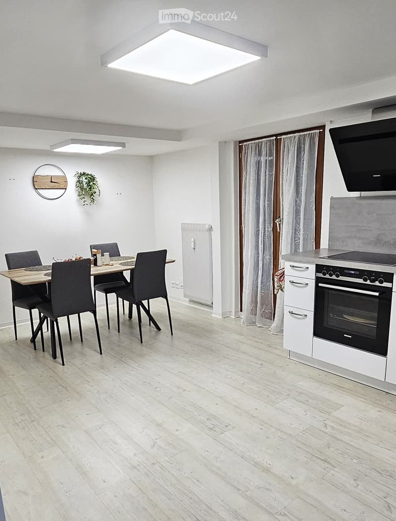 Open-plan kitchen and dining area with light wood flooring, white walls, and modern appliances including an oven and fridge. A dining table with four chairs is positioned near a window with curtains.