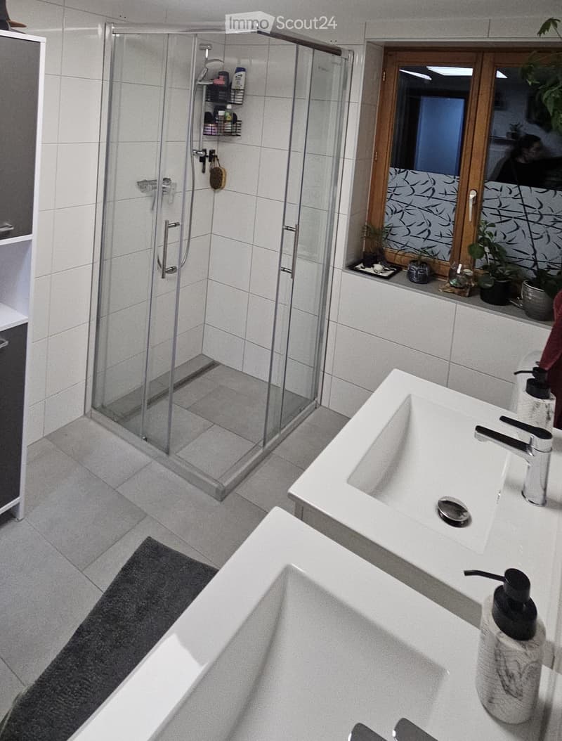 Modern bathroom with a large bathtub, double sink, glass-enclosed shower, and a window with plants on the sill.