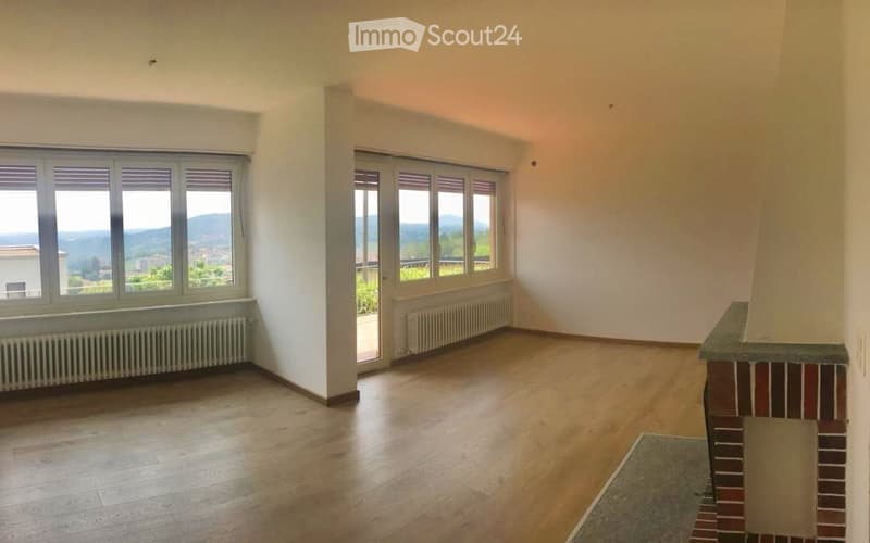 Empty room with wooden flooring, large windows offering a scenic view, and a fireplace base.