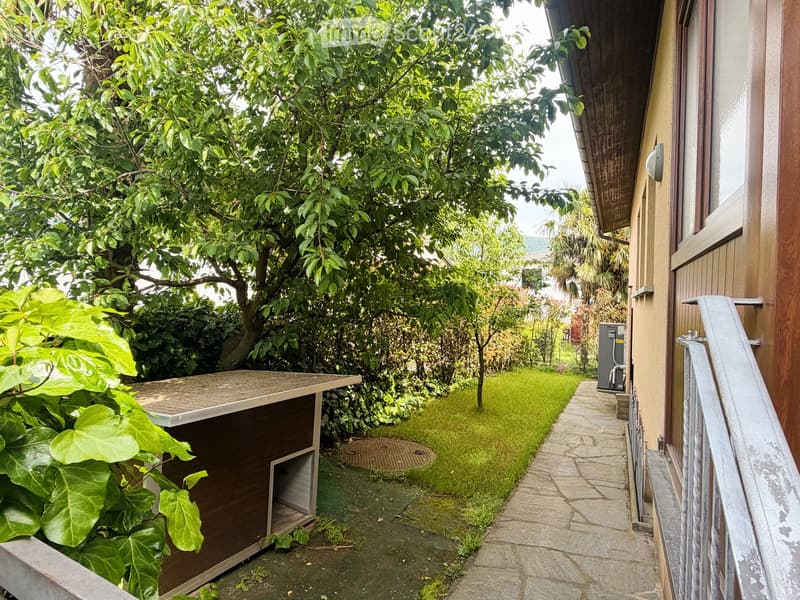 A small, well-maintained garden area with a wooden doghouse, a small table, lush greenery, and a paved pathway leading to the house.