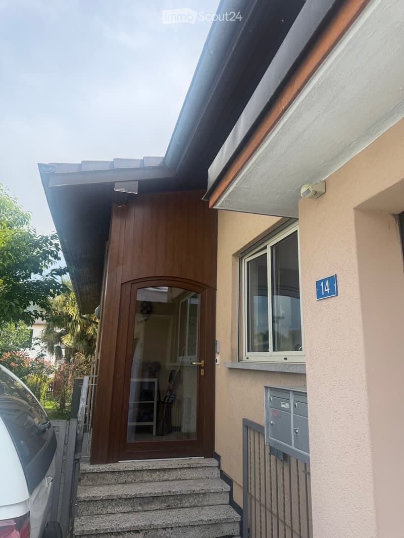 Front entrance of a two-story building with a wooden door, small steps, a window, and a mailbox.
