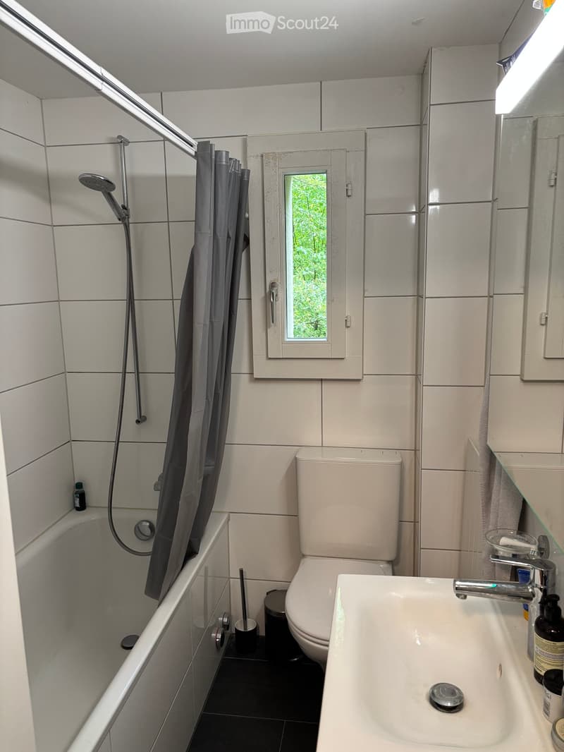 Modern bathroom with white tiles, bathtub, toilet, sink, shower, and a small window with a view of greenery.