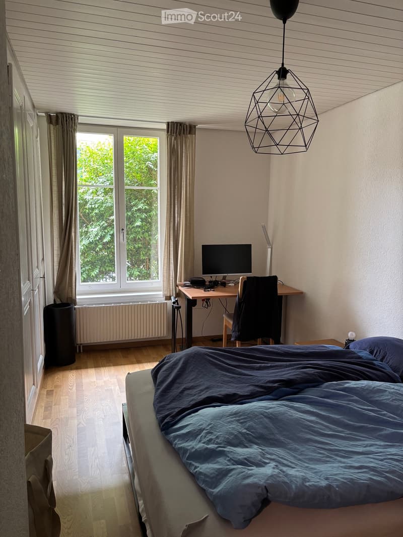 Cozy bedroom with a double bed, desk, chair, and computer monitor. Features wooden flooring, large window with curtains, and a modern geometric pendant light.