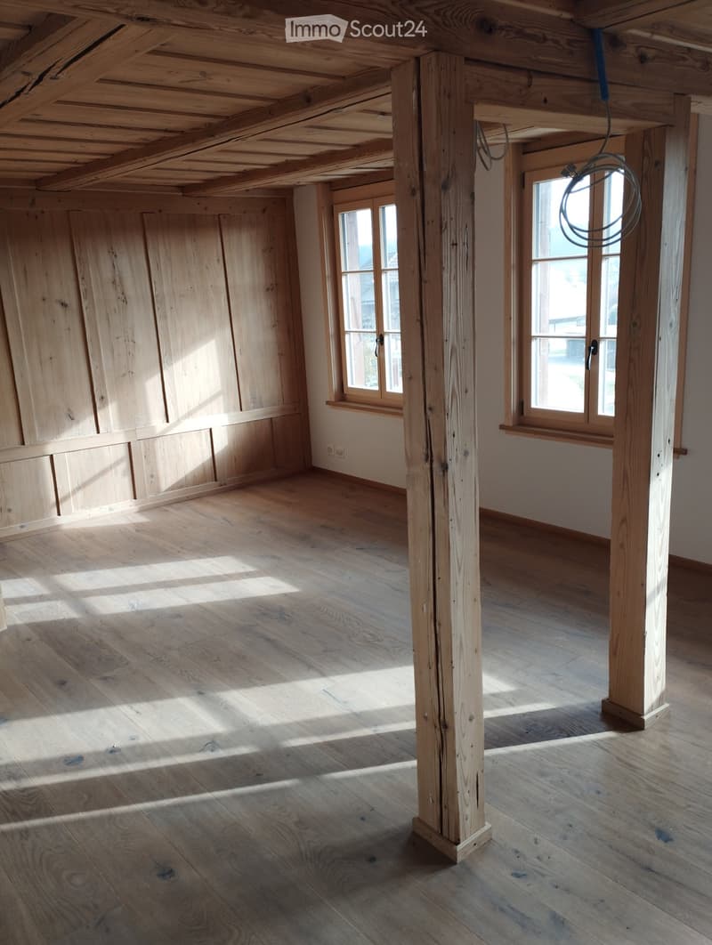 Empty room with wooden floor, exposed wooden beams, and large windows allowing natural light to enter. No furniture or equipment present.