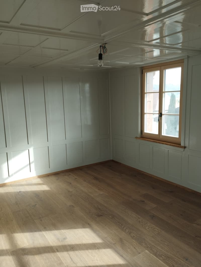 Empty room with light green walls, wooden floor, and large windows allowing natural light to enter. The ceiling features decorative molding and a skylight.