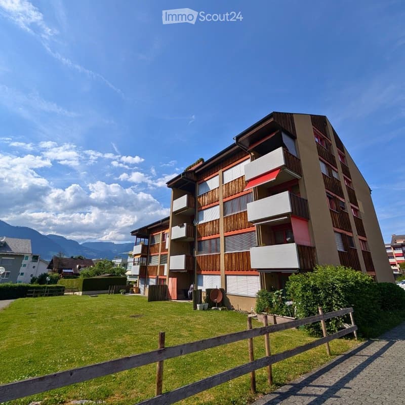 Modern multi-story residential building with balconies, wooden paneling, and a well-maintained lawn in front.