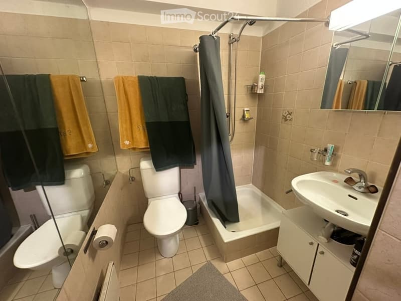 A small bathroom with beige tiled walls and floor, featuring a toilet, bathtub with a shower curtain, a sink with a mirror, and a towel rack with towels.