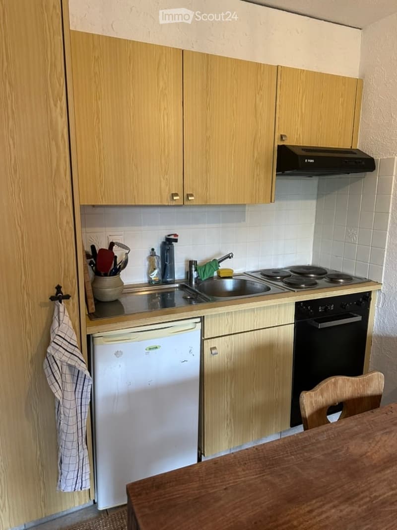 Compact kitchen with light wooden cabinets, a sink, stove, oven, and dishwasher. A small wooden table and chair are visible in the foreground.