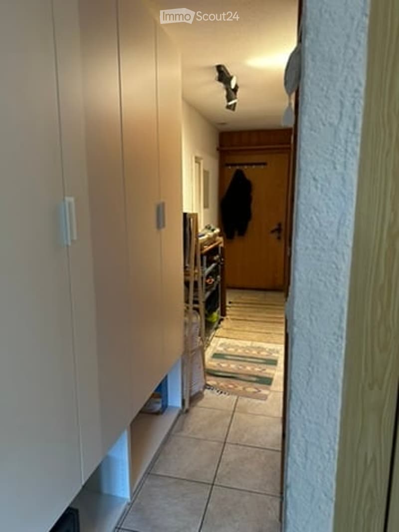 A narrow hallway with light-colored wooden cabinets on one side and a tiled floor. A small rug is placed near the end of the hallway, and a coat is hanging on a hook near a wooden door.