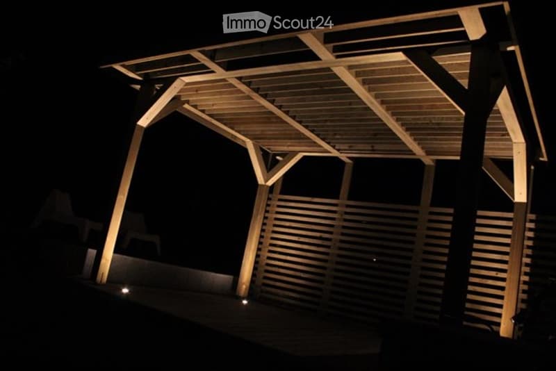 A wooden pergola structure illuminated by warm lighting, featuring a slatted roof and vertical wooden panels, set against a dark night sky.