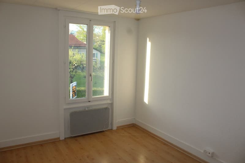 Empty room with wooden flooring, white walls, a window with a view of a garden, and a radiator below the window.