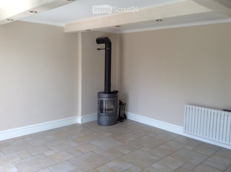 Empty room with tiled floor, beige walls, white baseboards, and a modern wood-burning stove with a chimney. A white radiator is mounted on the wall.