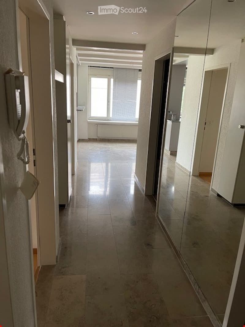 Empty hallway with tiled floor, white walls, and a large window at the end. Doors and a mirror are visible on the right side.