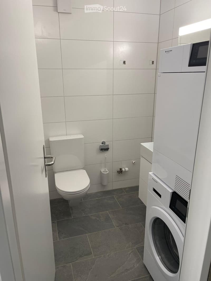 Modern bathroom with white tiled walls, a toilet, a washing machine, and a toilet paper holder.