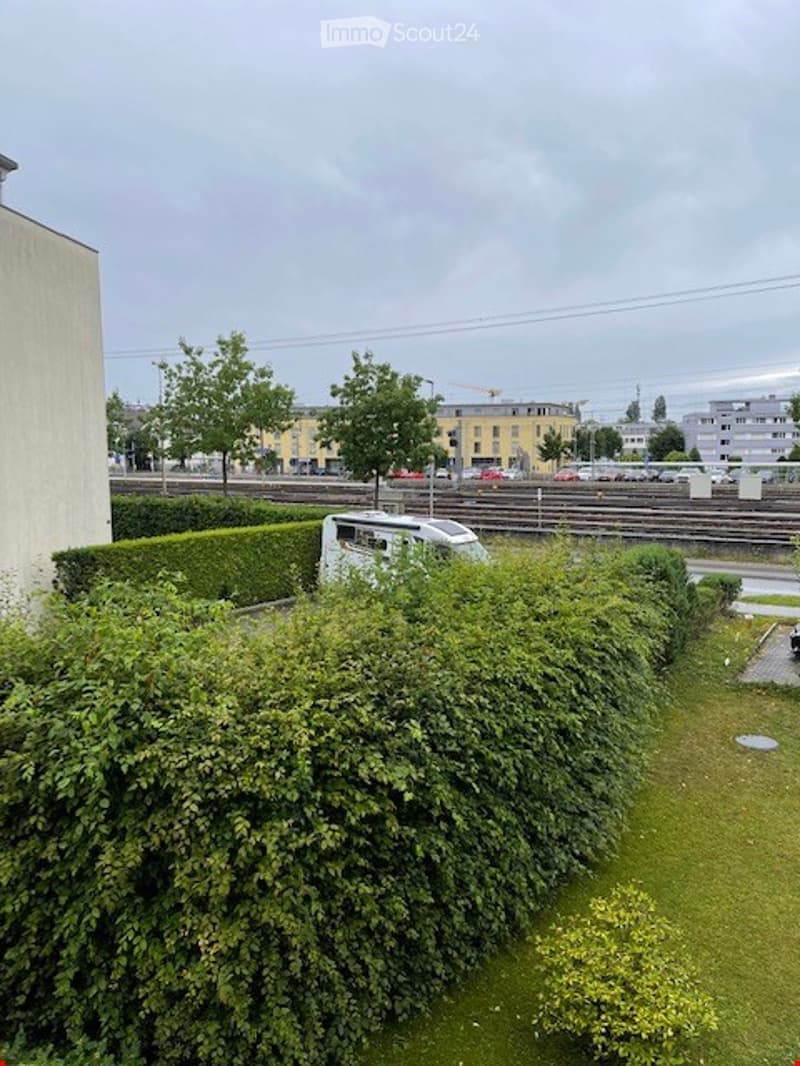 A small, enclosed garden area with neatly trimmed hedges, a small lawn, and a few trees. A white van is parked behind the hedge, and a railway track runs in the background.
