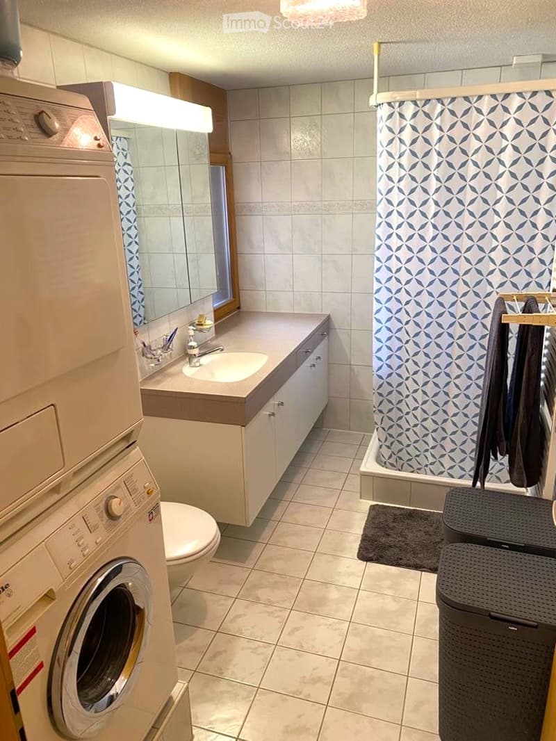 Modern bathroom with a sink, toilet, bathtub with a geometric shower curtain, and a private washing machine. Features include a large mirror, towel rack, and a small trash bin.