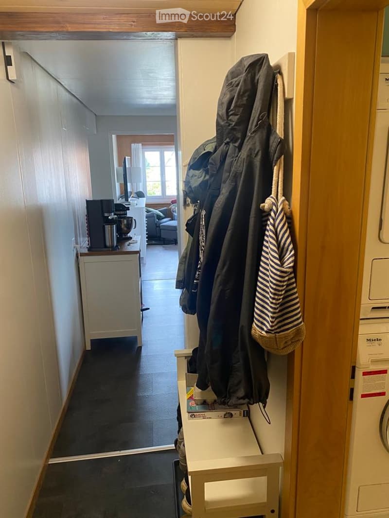 A narrow hallway with a coat rack holding jackets and a striped towel, leading to a living area with a sofa, coffee table, and a small kitchen counter with appliances.