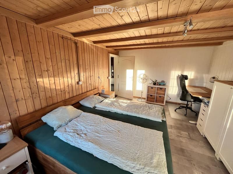 Cozy bedroom with wooden walls and ceiling, two single beds with green mattresses and white bedding, a desk with a chair, a fan, and a small storage unit.