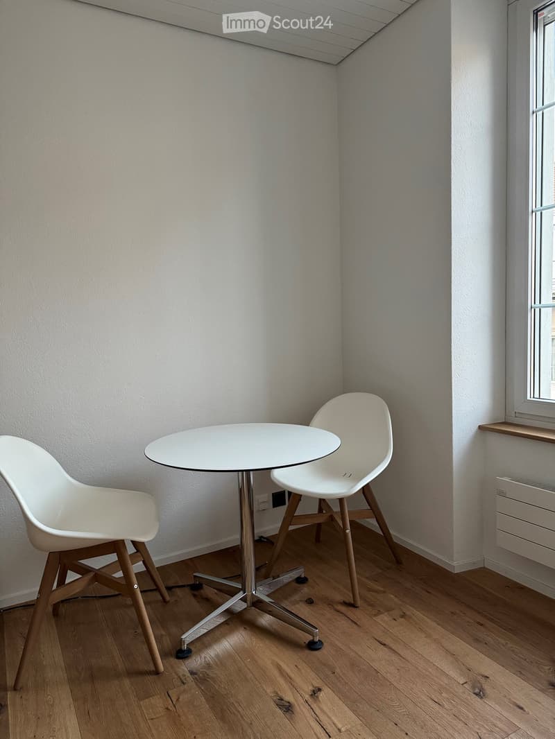 A small, modern room with light wooden flooring, white walls, and a large window. It features a round white table with a metal base and two white chairs with wooden legs.