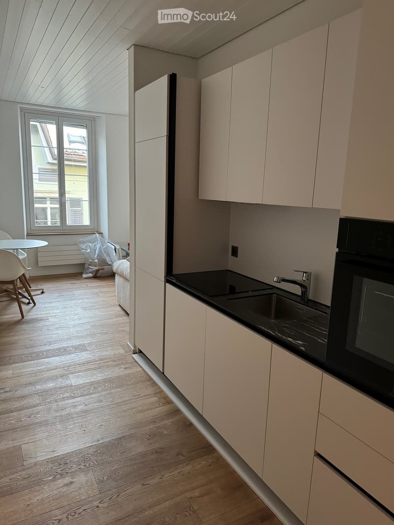 Open-plan kitchen and living area with light wood flooring, white cabinets, black countertop, sink, faucet, and a small table with a chair.