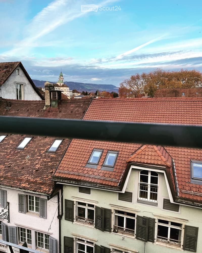 A multi-story residential building with red-tiled roofs, white walls, and multiple windows with shutters. The scene includes a distant church spire and mountains in the background.