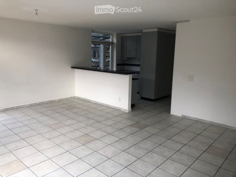 Empty room with tiled floor, white walls, and a small kitchen area with cabinets and a countertop.