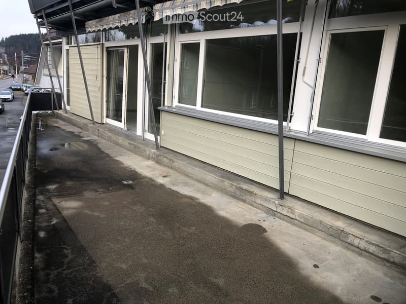 Balcony with a metal railing, wet concrete surface, and a view of a building with large windows and a covered entrance.