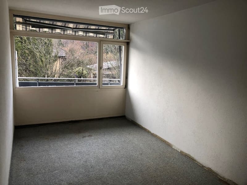 Empty room with carpeted floor, white walls, and large window with a view of trees and neighboring buildings.