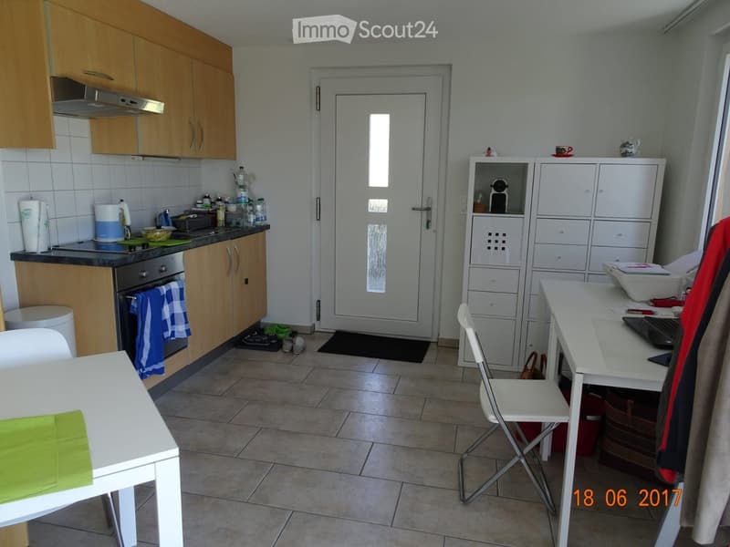 The image shows a modern kitchen with wooden cabinets, a stove, oven, and refrigerator. There is a dining table with a green napkin, a white chair, and a desk with a laptop and other items. The floor is tiled, and there is a door leading outside.