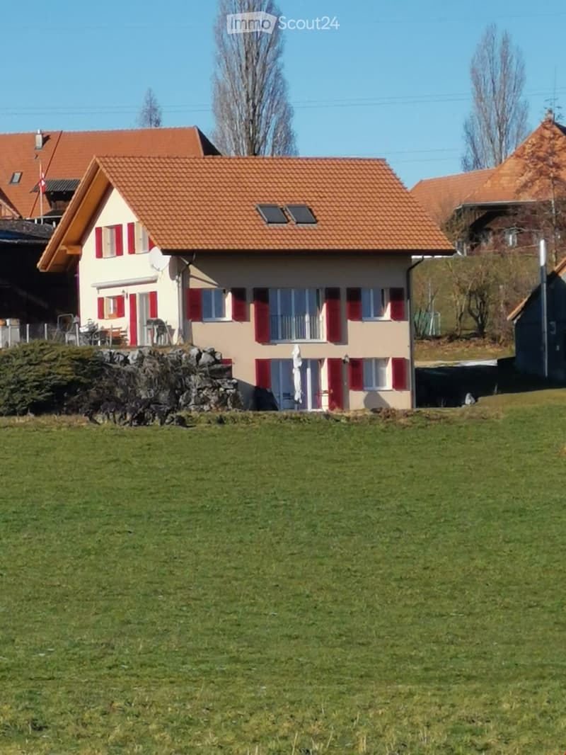 Two-story house with red-tiled roof, beige walls, red shutters, and a small balcony. The house is surrounded by a grassy lawn and a stone wall.