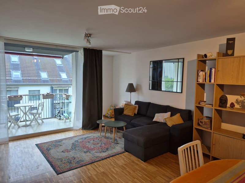 Modern living room with a dark sectional sofa, coffee table, side table, floor lamp, bookshelf, and a balcony with outdoor furniture.