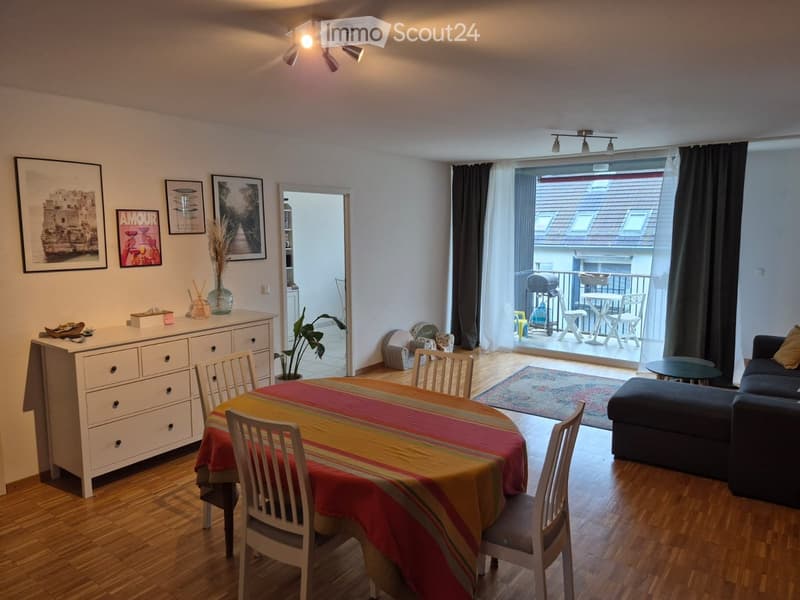 Bright living and dining area with wooden flooring, a sofa, ottoman, coffee table, dining table with chairs, and a balcony leading to an outdoor space.