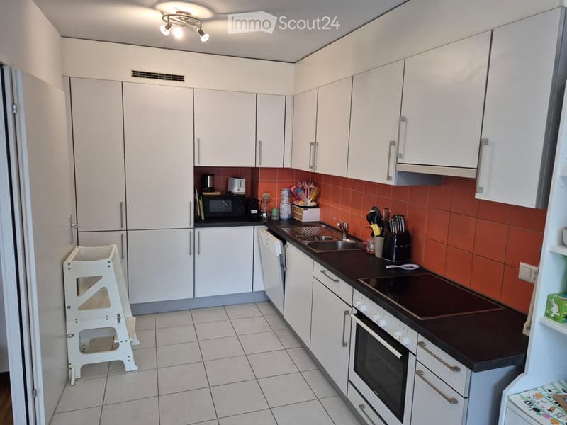 Modern kitchen with white cabinets, dark countertops, and stainless steel appliances including an oven, microwave, and sink. Features a tiled backsplash in orange and a small white stool near the fridge.