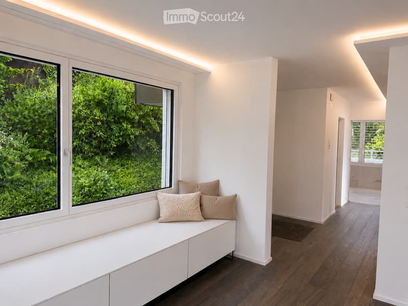 Modern, minimalist room with a built-in bench and decorative pillows, large windows with lush greenery view, wooden flooring, and recessed lighting.