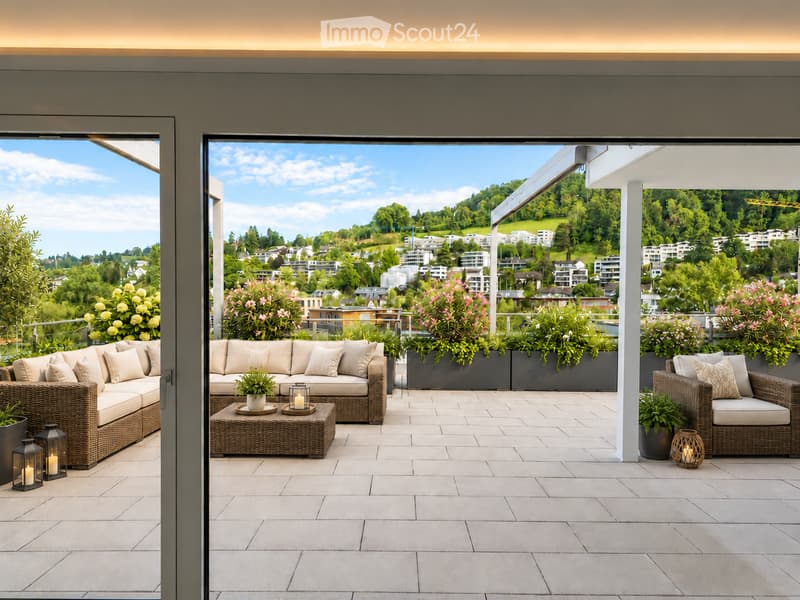 A spacious balcony with a modern design, featuring a wicker sofa, coffee table, chairs, lanterns, and potted plants, overlooking a scenic cityscape with greenery.