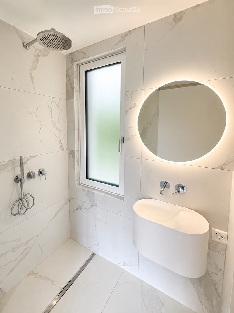 Modern bathroom with marble tiles, a glass shower enclosure, a sleek white sink, and a round illuminated mirror.