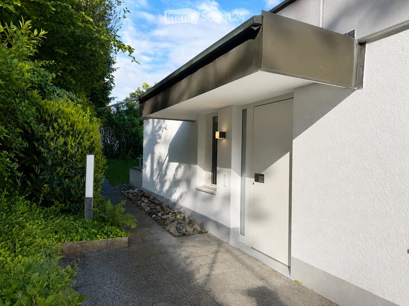 Modern white building with a sleek, minimalist design, featuring a covered entrance with a metallic roof overhang and a small window. The surrounding area includes a well-maintained garden with lush greenery and a stone pathway leading to the entrance.