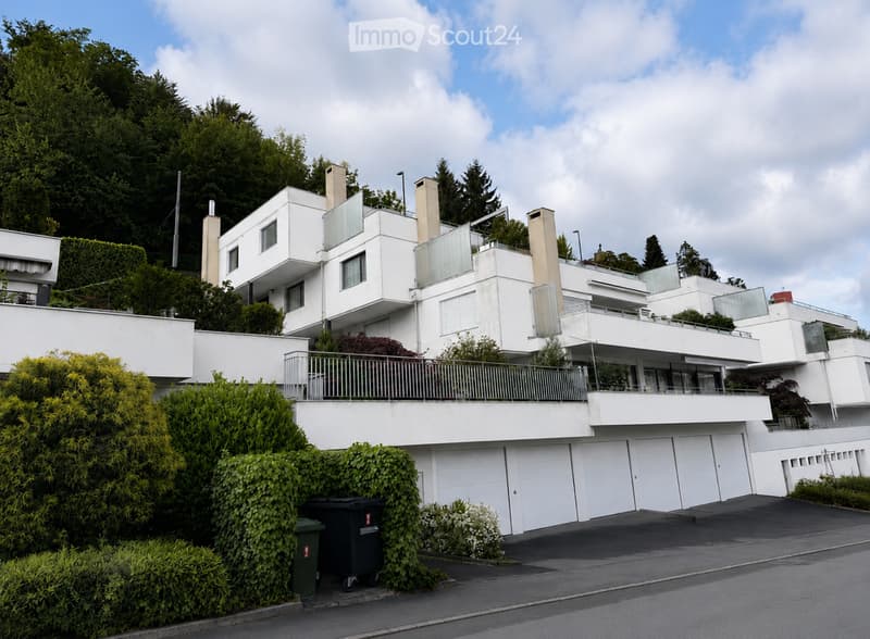 Modern white multi-level house with large windows, balconies, and a garage, surrounded by greenery.