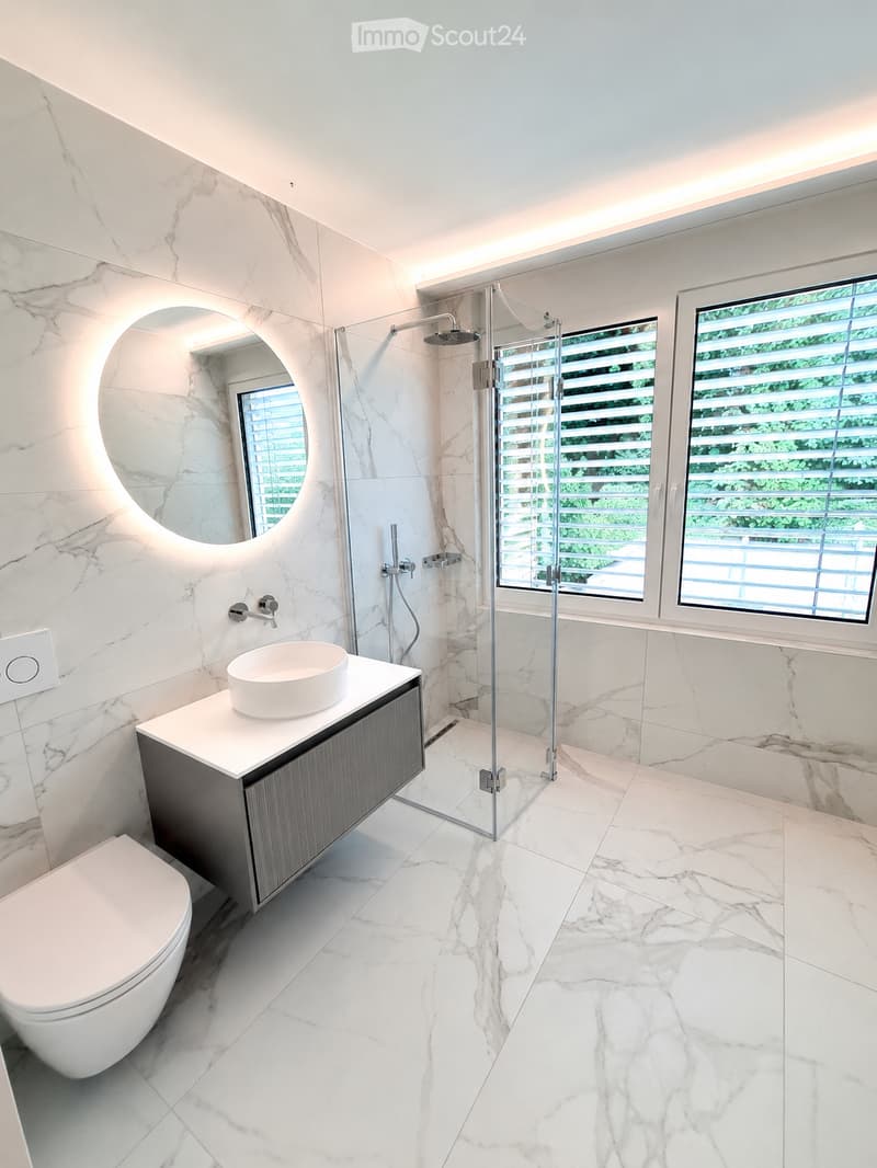 Modern bathroom with white marble tiles, a toilet, a sink with a modern faucet, a large round mirror, and a glass-enclosed shower with a modern showerhead.