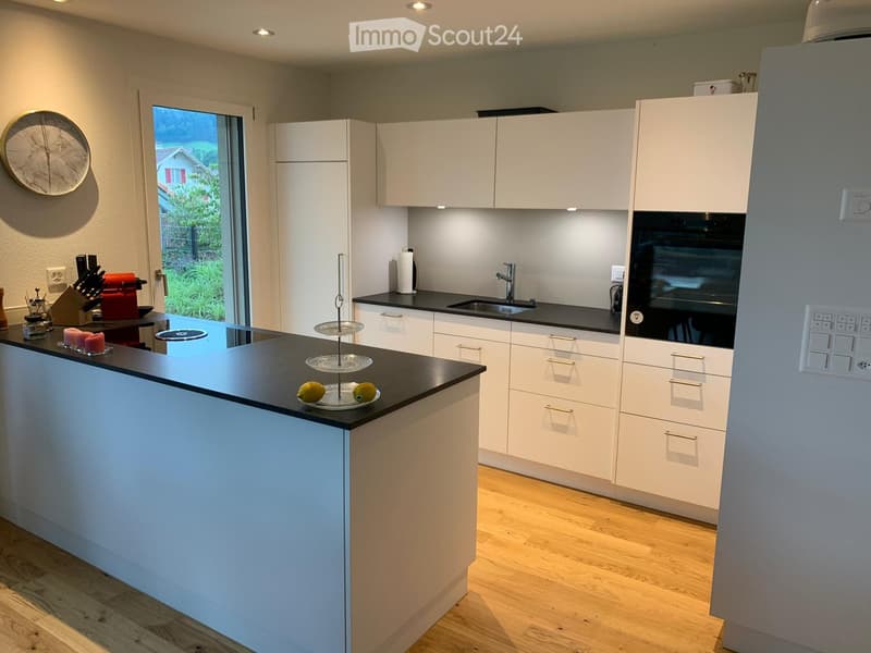 Modern kitchen with white cabinets, black countertops, and a central island. Features include a sink, oven, coffee maker, knife block, and decorative cake on a stand.