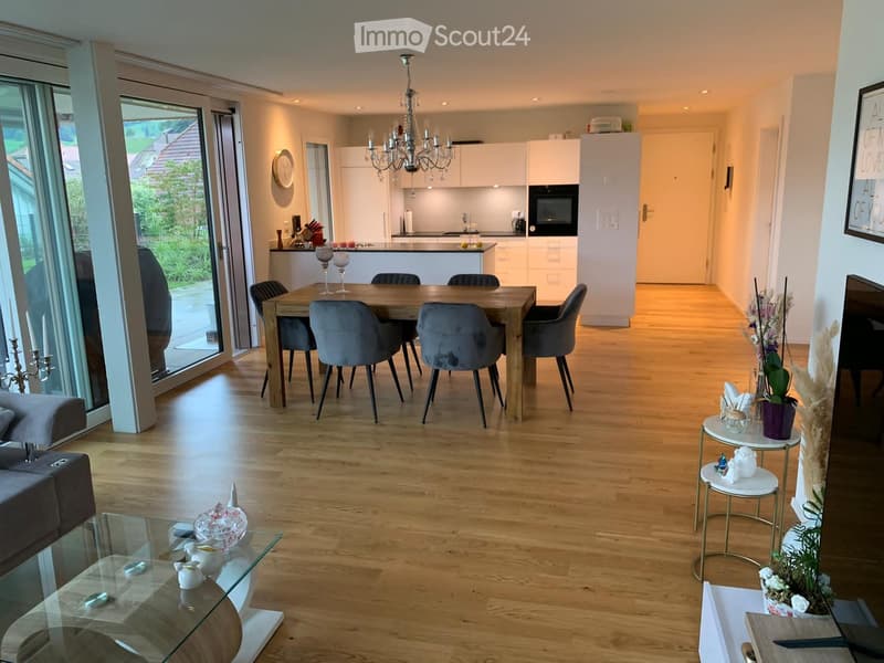 Open-plan living and dining area with light wooden flooring, a modern sofa, a glass coffee table, a dining table with six chairs, and a fully equipped kitchen with white cabinets and a microwave. The space features a chandelier, side tables with decorativ