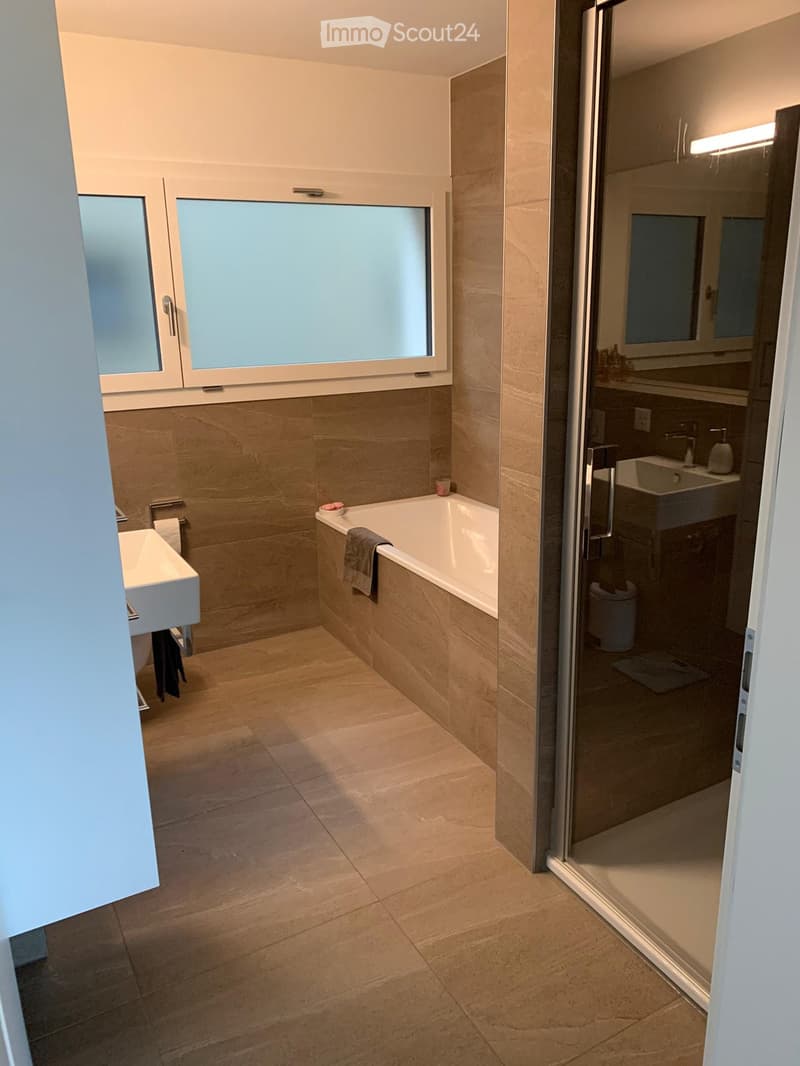 Modern bathroom with beige tiles, a bathtub, two sinks, a large mirror, and a glass shower enclosure.