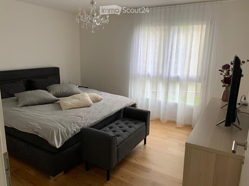 Modern bedroom with a large bed, bench, TV, chandelier, and flower vase on a side table.